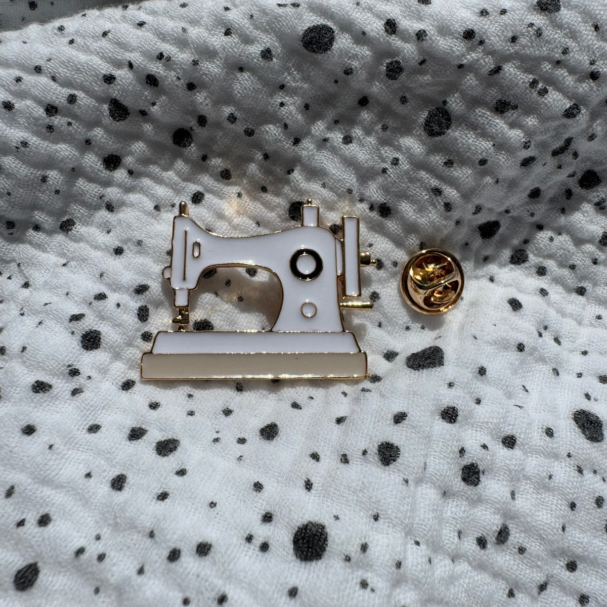 Emaille-Pin „Nähmaschine“ auf gepunktetem Stoff, daneben der Pin-Verschluss