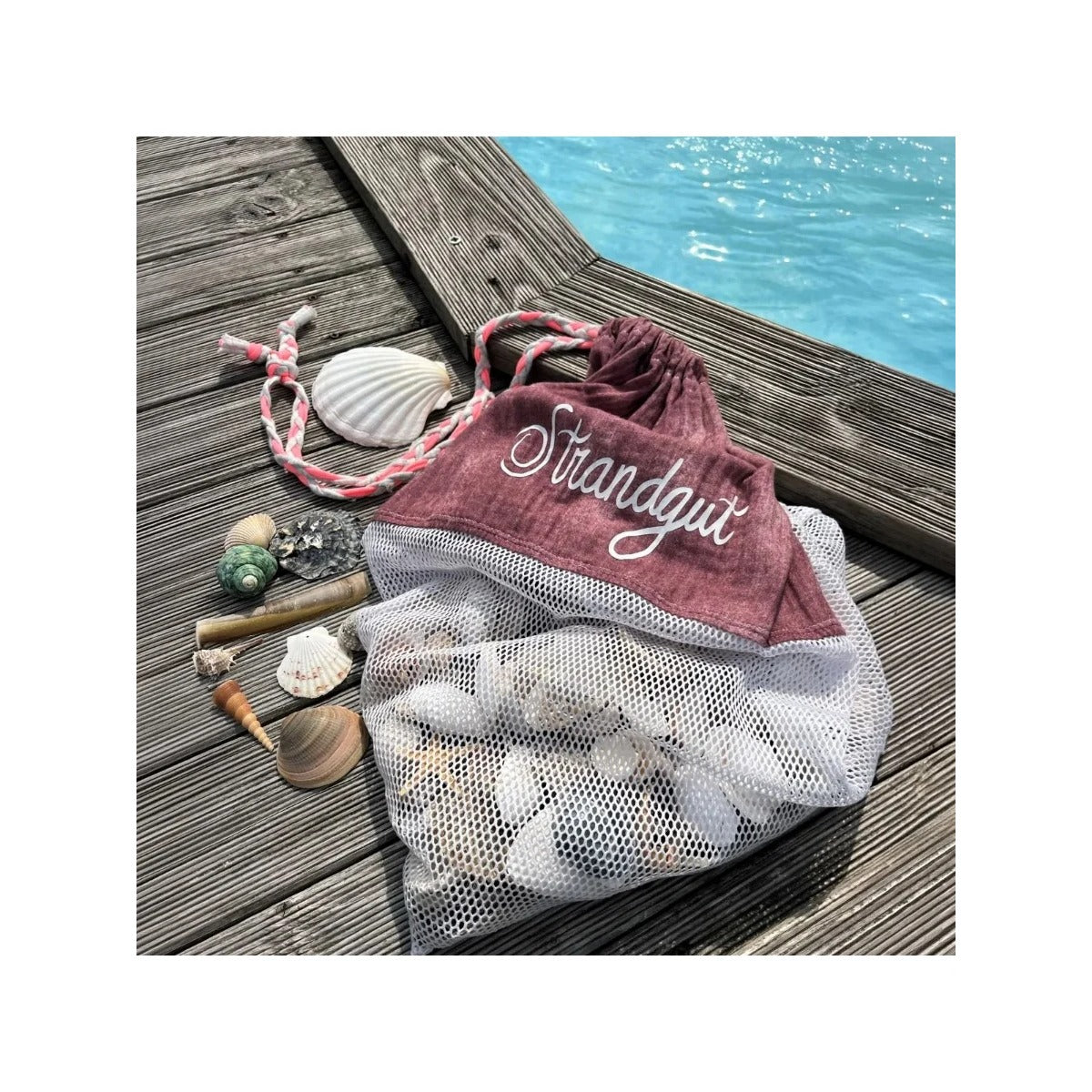 Freebook Sammelbeutel Strandgut – beerenfarbener Netzbeutel mit Schriftzug Strandgut (aus der passenden Plotterdateiserie mit diesem Namen), gefüllt mit Muscheln, auf einem Steg am Wasser