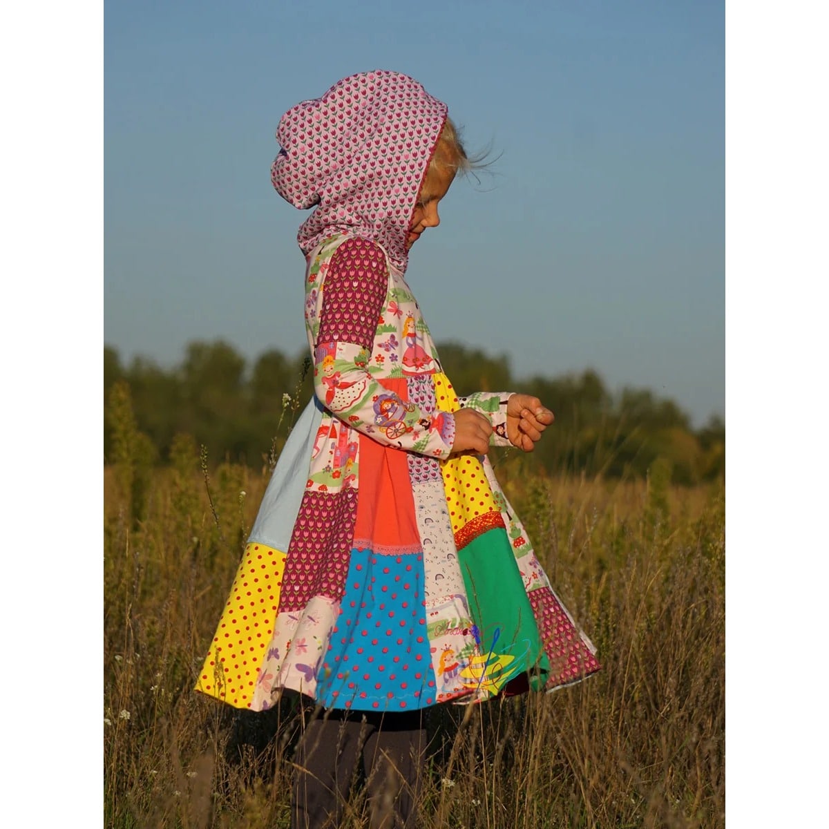 Lottis Drehkleid Schnittmuster – Mädchen im Feld trägt Drehkleid mit Kapuze und buntem Patchwork-Rock aus Prinzessinnen-, Punkte- und Tulpenstoffen