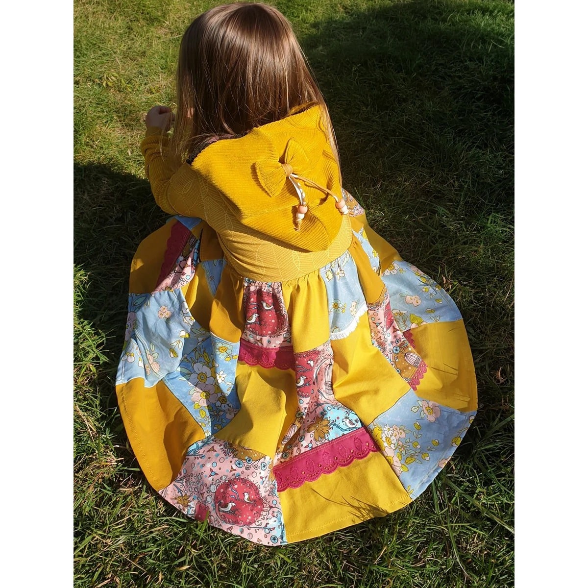 Lottis Drehkleid Schnittmuster – sitzendes Mädchen im senfgelben Kleid mit Stretchcord-Kapuze, Schleife und Patchworkrock mit eingenähten Spitzen