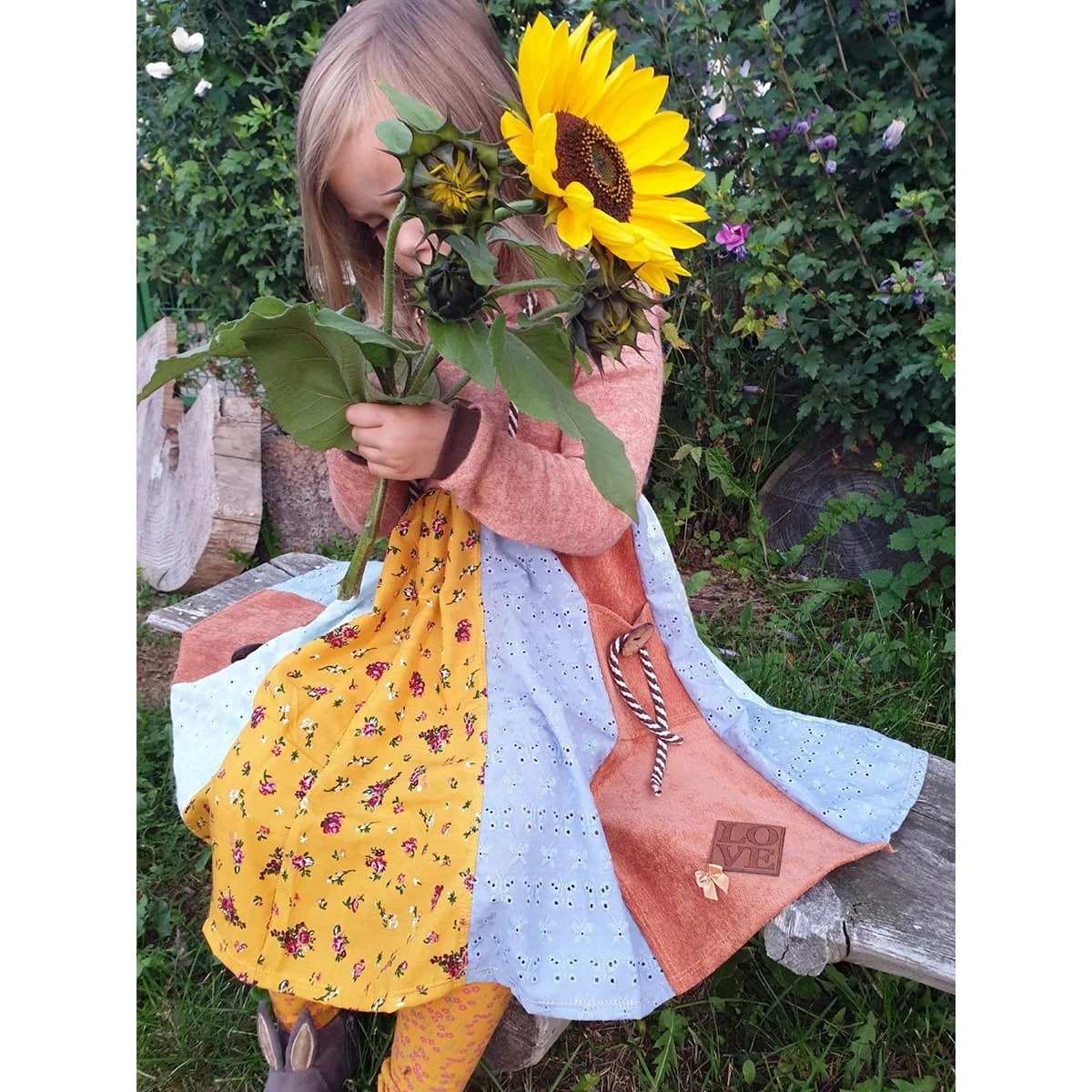 Lottis Drehkleid Schnittmuster – Mädchen mit Sonnenblumen, koralles Oberteil aus Sommersweat und Rock mit Blümchen, blauer Lochspitze und LOVE-Label