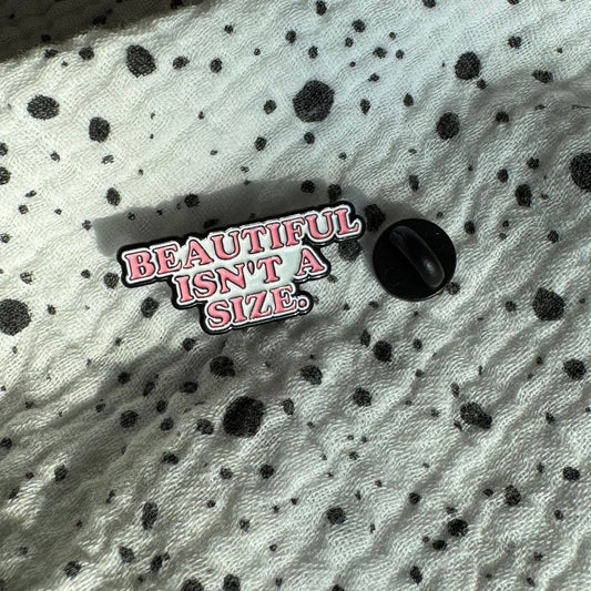 Emaille-Pin „Beautiful isn’t a size“ auf gepunktetem Stoff, daneben der Pin-Verschluss