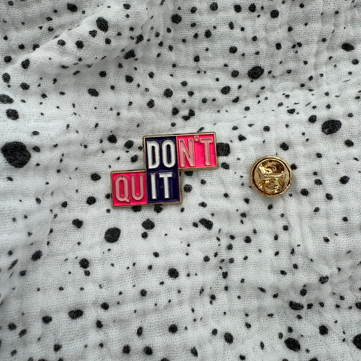 Emaille-Pin „Don’t quit – DO IT“ auf gepunktetem Stoff, daneben der Pin-Verschluss