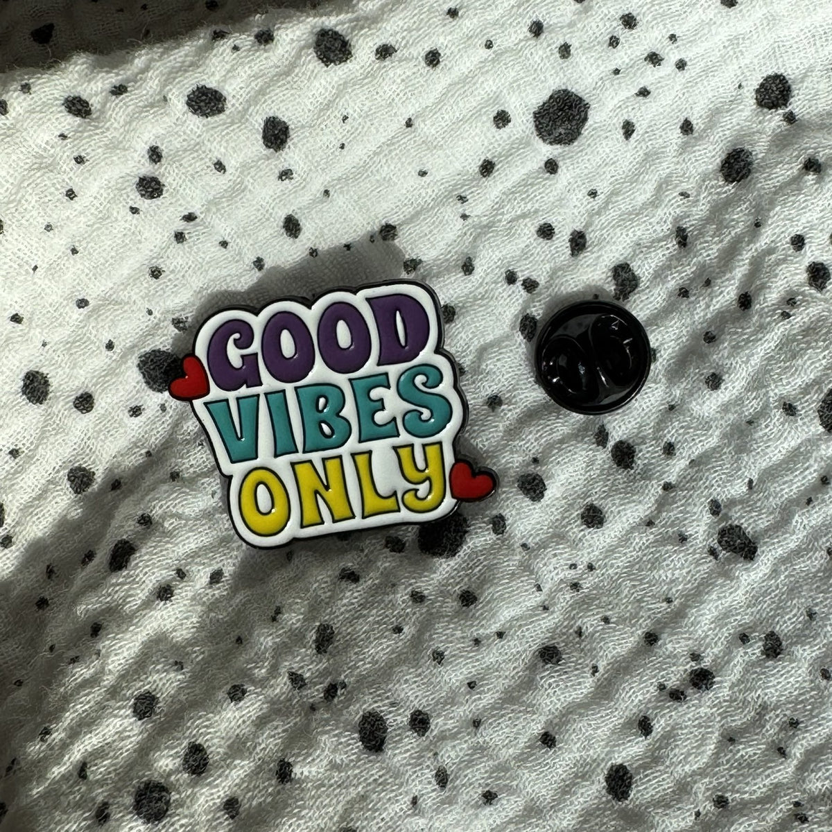 Emaille-Pin „Good Vibes Only“ auf gepunktetem Stoff, daneben der Pin-Verschluss
