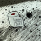 Emaille-Pin „I don’t give a shit“ auf gepunktetem Stoff, daneben der Pin-Verschluss