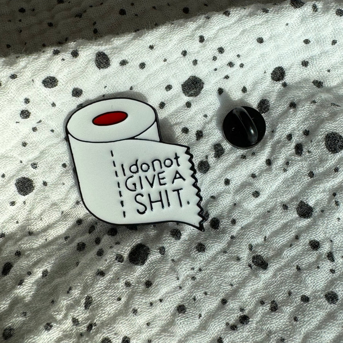Emaille-Pin „I don’t give a shit“ auf gepunktetem Stoff, daneben der Pin-Verschluss