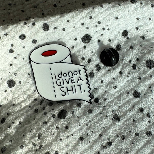 Emaille-Pin „I don’t give a shit“ auf gepunktetem Stoff, daneben der Pin-Verschluss