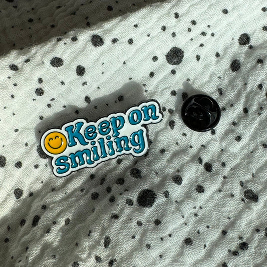 Emaille-Pin „keep smiling“ auf gepunktetem Stoff, daneben der Pin-Verschluss