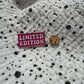 Emaille-Pin „Limited Edition“ auf gepunktetem Stoff, daneben der Pin-Verschluss