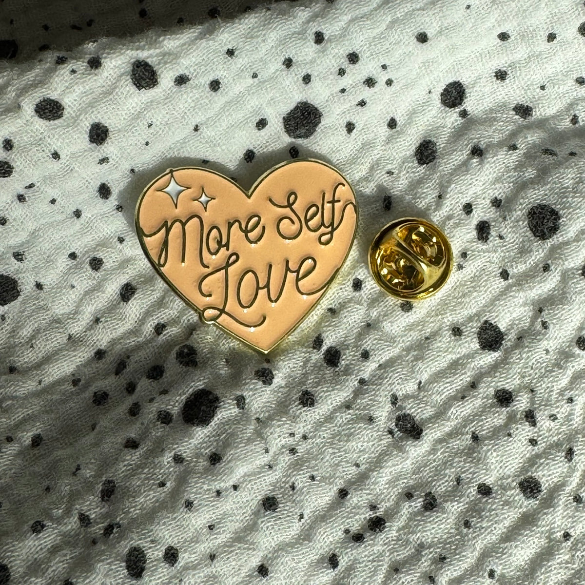 Emaille-Pin „More Self Love“ auf gepunktetem Stoff, daneben der Pin-Verschluss