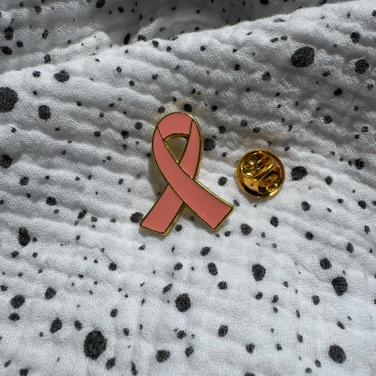 Emaille-Pin „Pink Ribbon“ gold auf gepunktetem Stoff, daneben der Pin-Verschluss