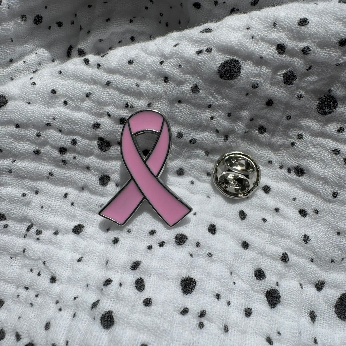 Emaille-Pin „Pink Ribbon“ silber auf gepunktetem Stoff, daneben der Pin-Verschluss