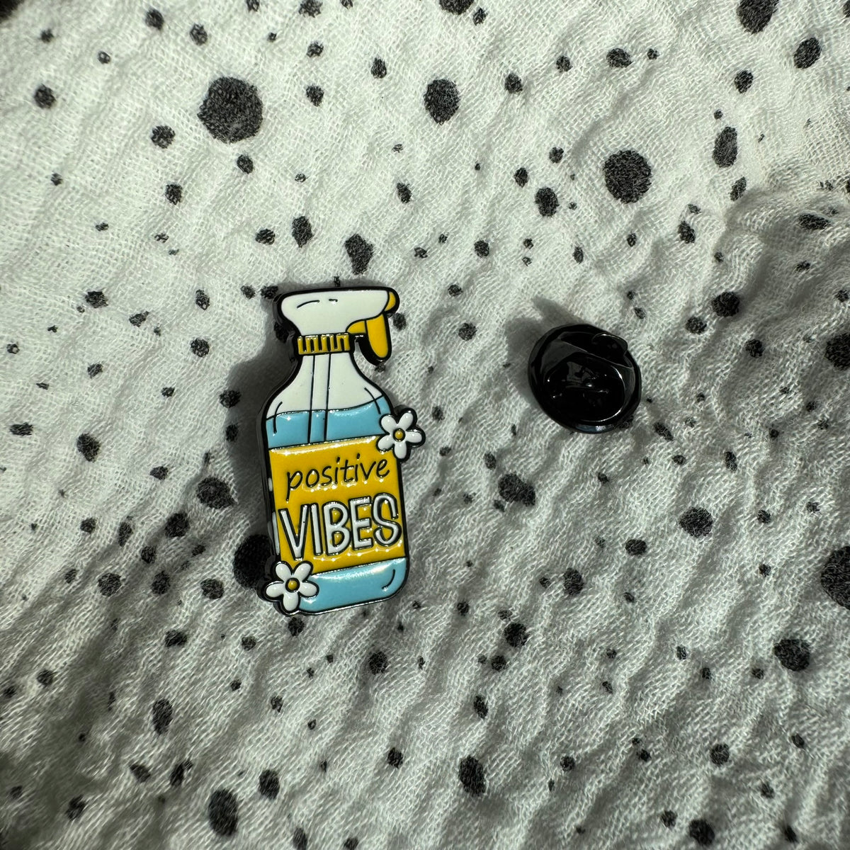 Emaille-Pin „Positive Vibes“ auf gepunktetem Stoff, daneben der Pin-Verschluss