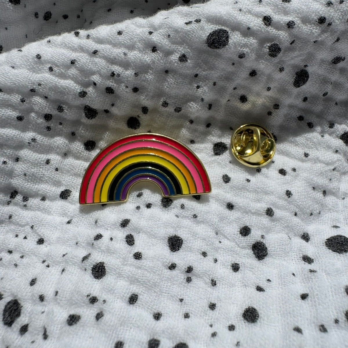Emaille-Pin „Regenbogen“ auf gepunktetem Stoff, daneben der Pin-Verschluss