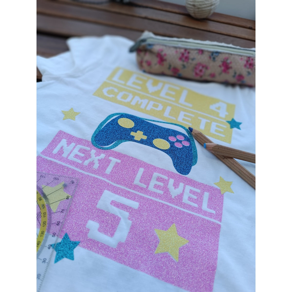 Gaming Plotterdatei – weißes Shirt mit den Motiven „Level 4 Complete“ und „Next Level 5“ in gelber und rosa Glitzerfolie, dazu ein blauer Glitzer-Controller und Sterne, als Idee zum Schulwechsel in die fünfte Klasse