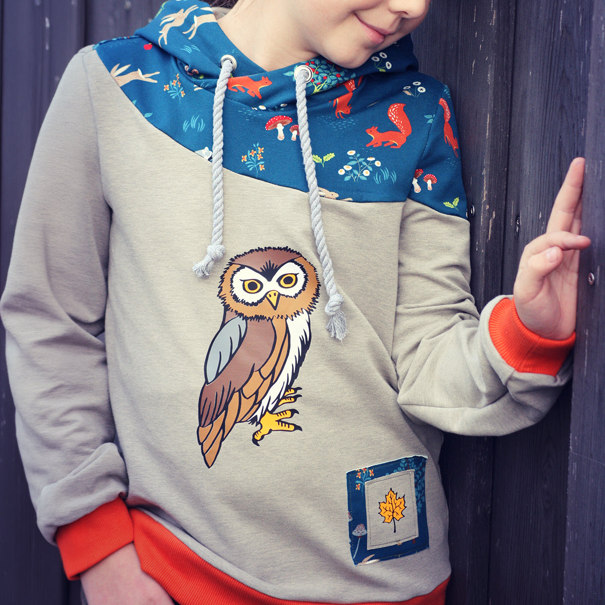 Herbstgrüße Plotterdatei – Eule auf beigem Hoodie mit blauer Waldtier-Passe, roten Bündchen und aufgesetzter Tasche mit Ahornblatt