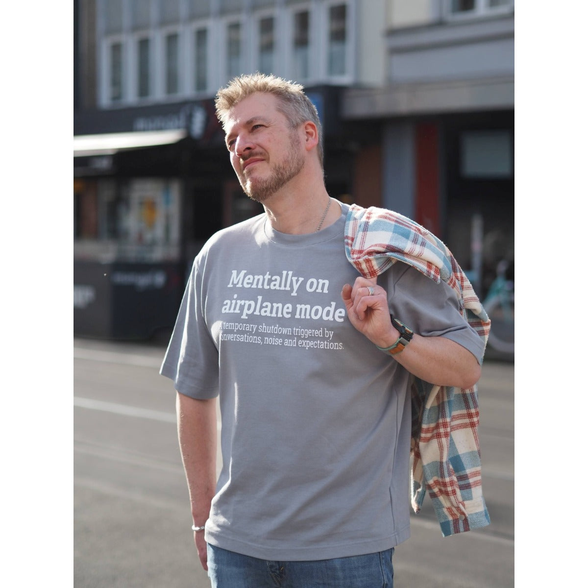 Intolerant Club Plotterdatei – Statement „Mentally on airplane mode“ weiß geplottet auf einem grauen Oversized-Shirt, Mann mit Holzfällerhemd über der Schulter