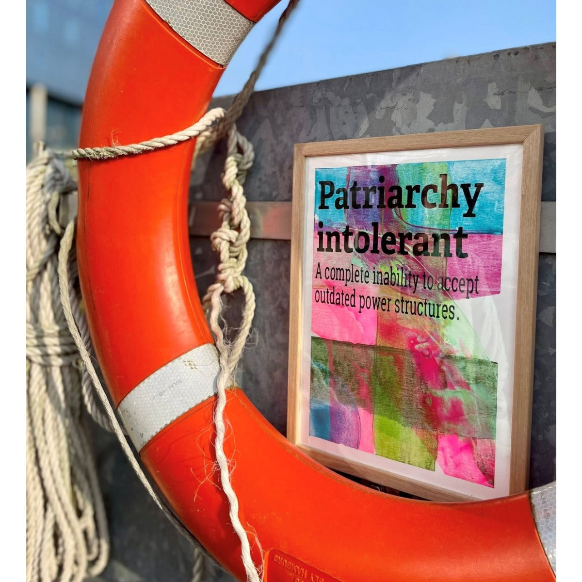 Intolerant Club Plotterdatei – Statement „Patriarchy intolerant“ schwarz auf buntem Gelli-Print, gerahmt als Bild und an einen orangefarbenen Rettungsring gelehnt