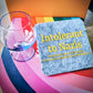 Intolerant Club Plotterdatei – Statement „Intolerant to Nazis“ in Gelb auf einem grauen Filz-Untersetzer, daneben ein Glas auf einem Regenbogen-Buch