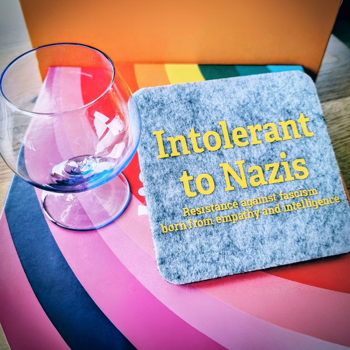 Intolerant Club Plotterdatei – Statement „Intolerant to Nazis“ in Gelb auf einem grauen Filz-Untersetzer, daneben ein Glas auf einem Regenbogen-Buch