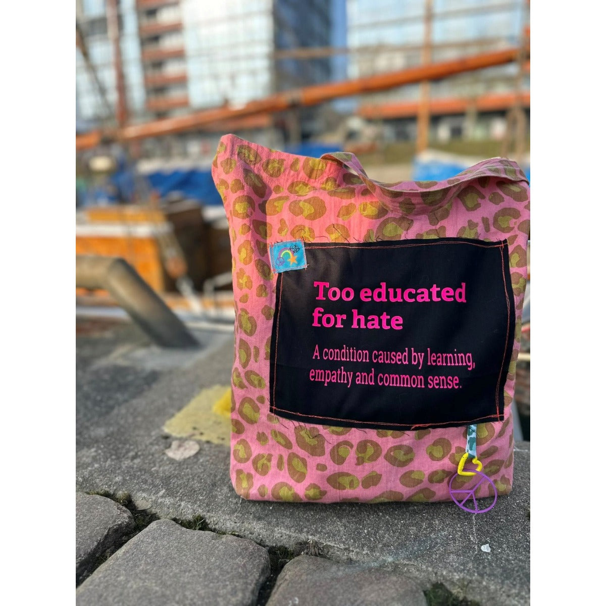 Intolerant Club Plotterdatei – Statement „Too educated for hate“ in Pink auf einem schwarzen Stoffrechteck, aufgenäht auf eine rosa Leo-Tasche mit Peace-Anhänger
