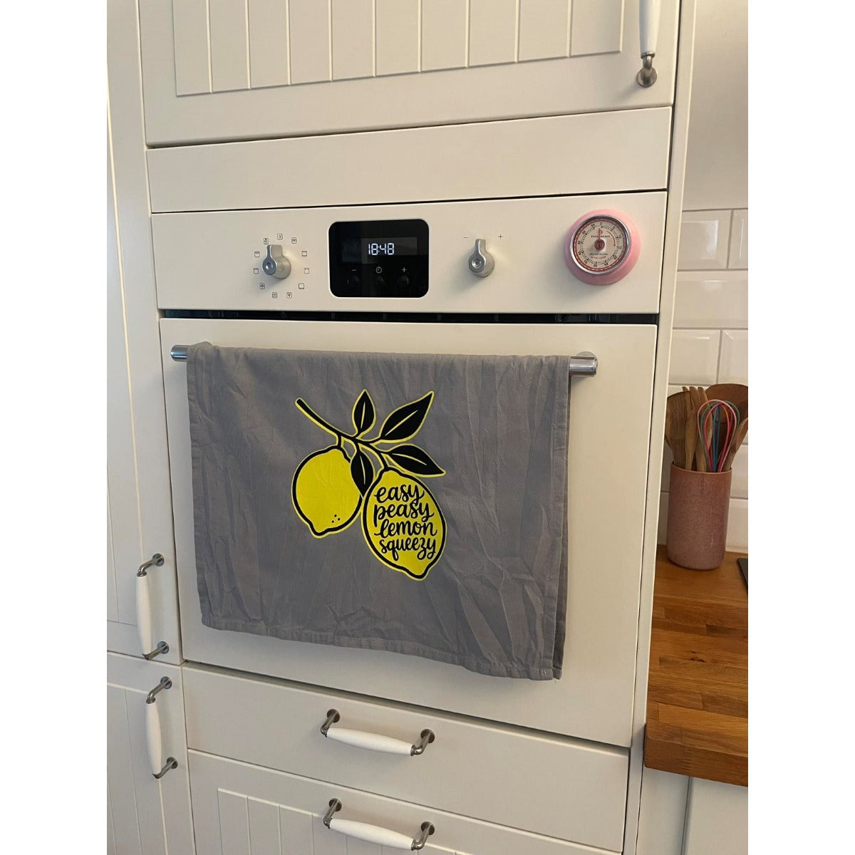 Graues Geschirrtuch mit Zitronenmotiv und Schriftzug Easy Peasy Lemon Squeezy, an der Küchenzeile hängend