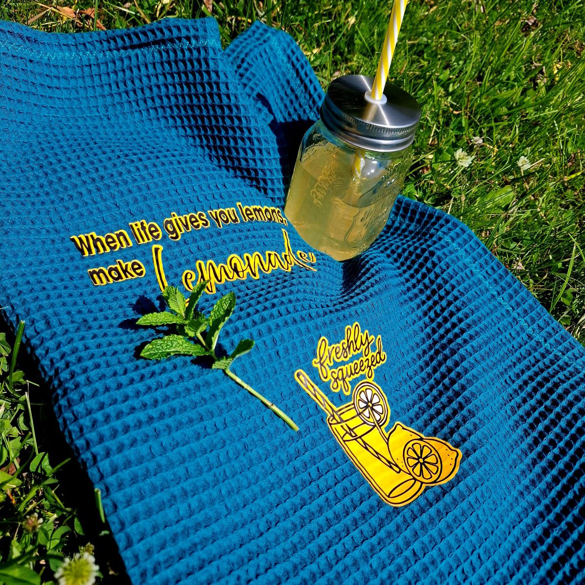 Blaues Waffelpiqué-Handtuch mit den Motiven When life gives you lemons, make Lemonade und Freshly Squeezed, daneben ein Trinkglas mit Minze
