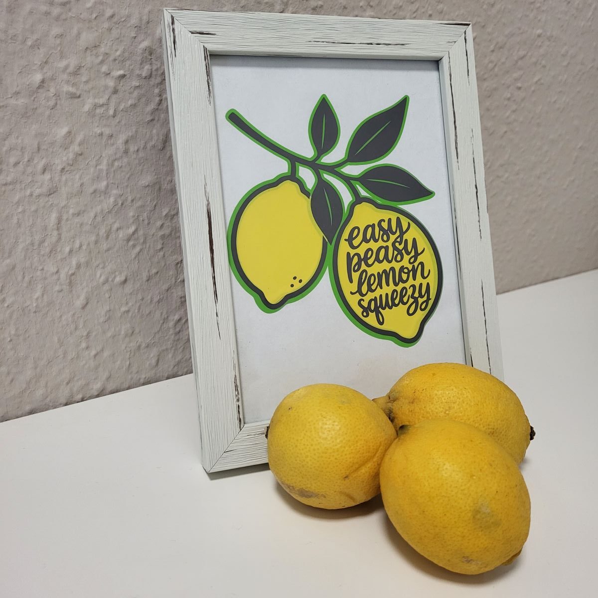 Weißer Bilderrahmen mit Zitronen-Layer-Motiv und Schriftzug Easy Peasy Lemon Squeezy, davor drei frische Zitronen