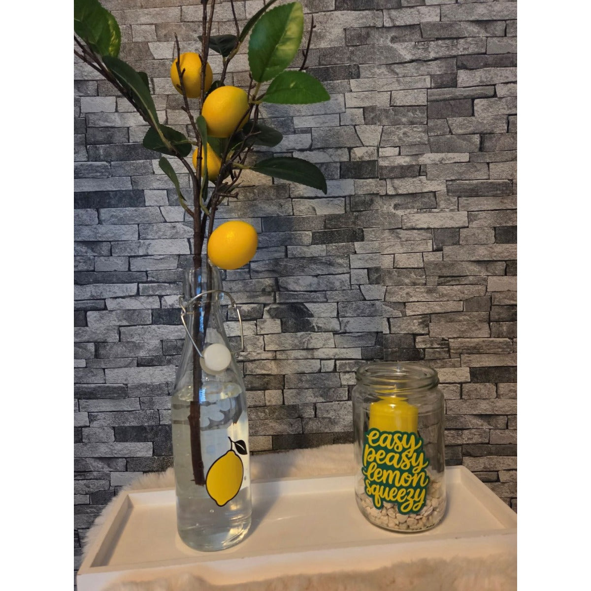 Glasflasche mit Zitronenmotiv und Windlicht mit dem Schriftzug Easy Peasy Lemon Squeezy vor einer Steinwand