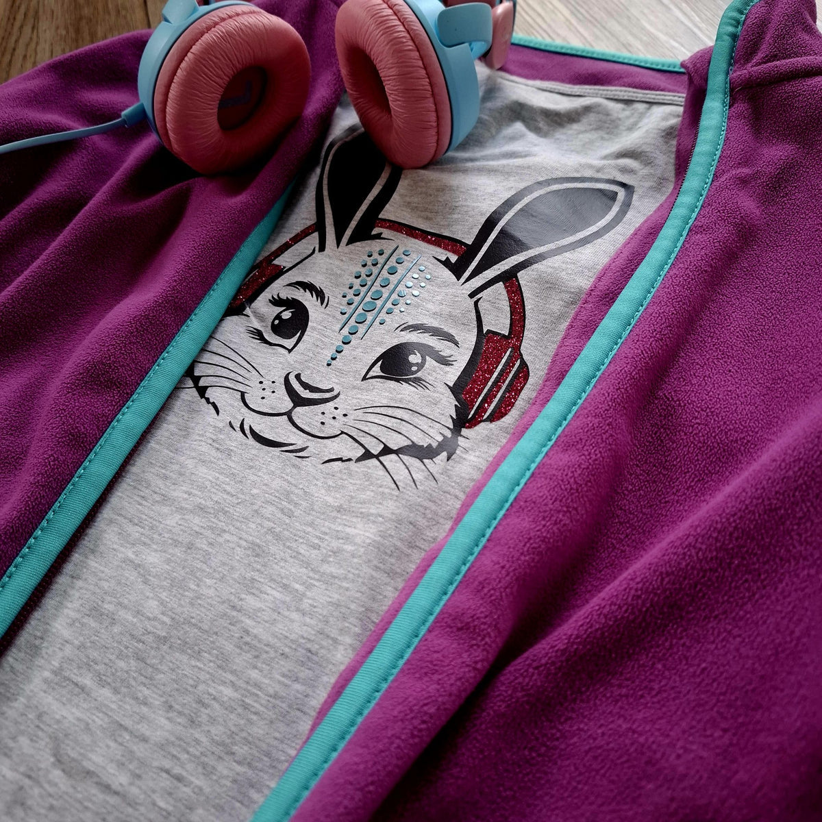 Grauer Hoodie mit Hasenmotiv in Glitzerfolie und pinkem Innenfutter, daneben echte Kopfhörer