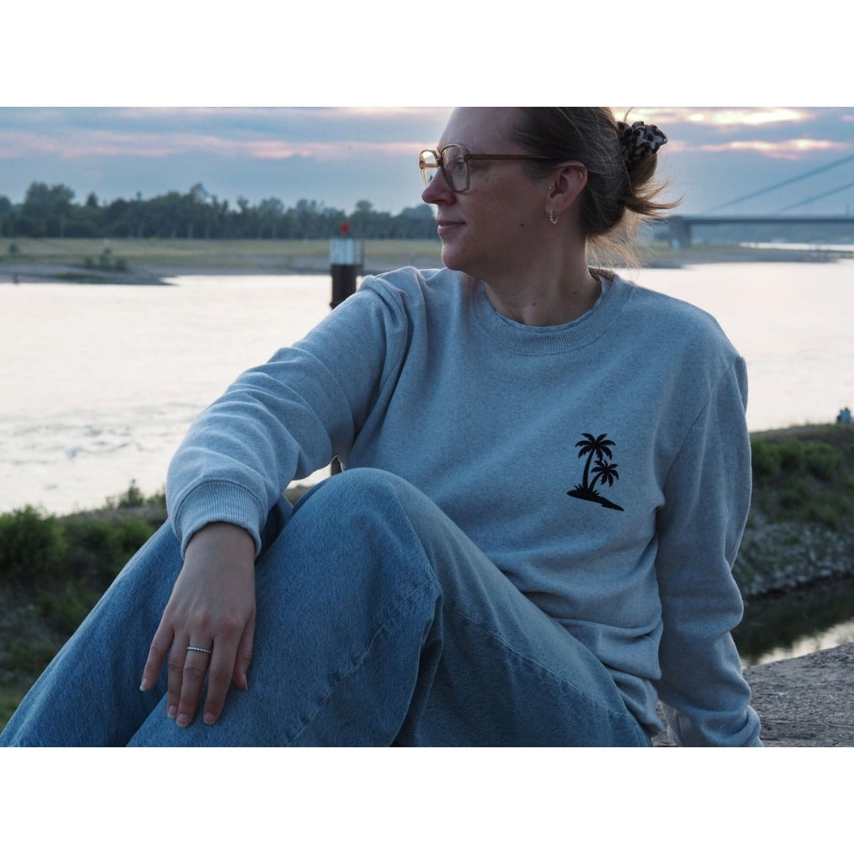 Frau im grauen Sweatshirt mit kleiner Palmen-Plotterdatei auf der Brust am Flussufer