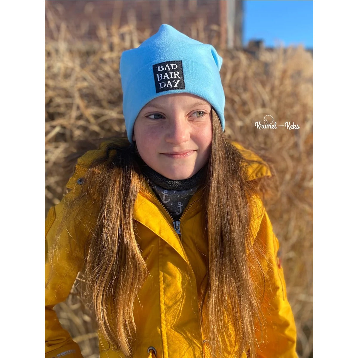 Beanie Bernie – hellblaue Umschlagbeanie mit ‚Bad Hair Day'-Label, getragen von einem Mädchen in gelber Jacke