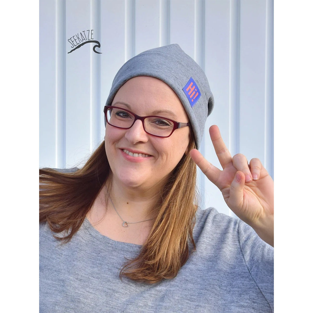 Beanie Bernie – graue Umschlagbeanie mit buntem ‚Hi!'-Label, getragen von einer Frau mit Brille