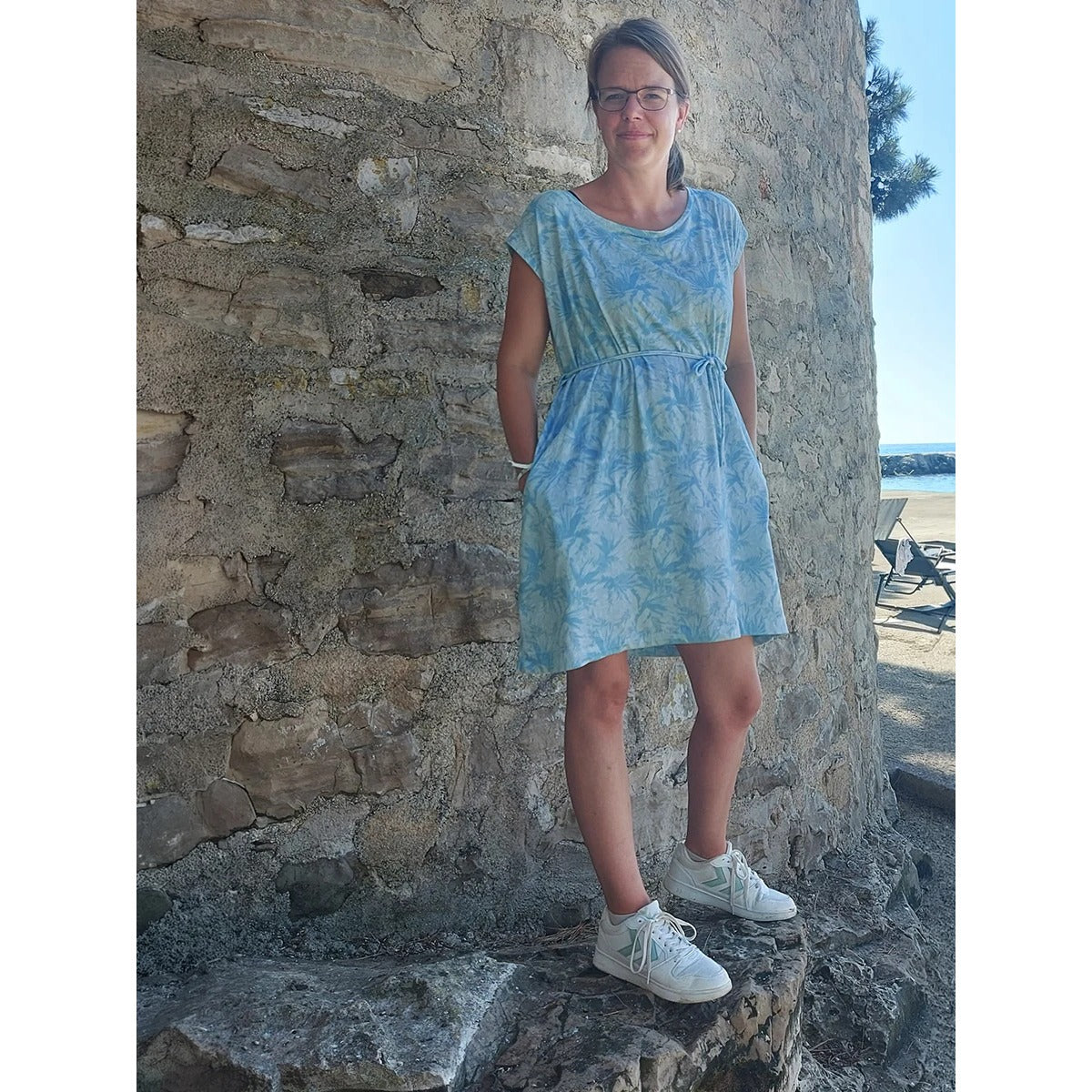 Cut Out Kleid – türkis gemustertes Kleid mit kurzen Ärmeln, Eingrifftaschen und Bindeband an der Hüfte, vor einer Steinwand
