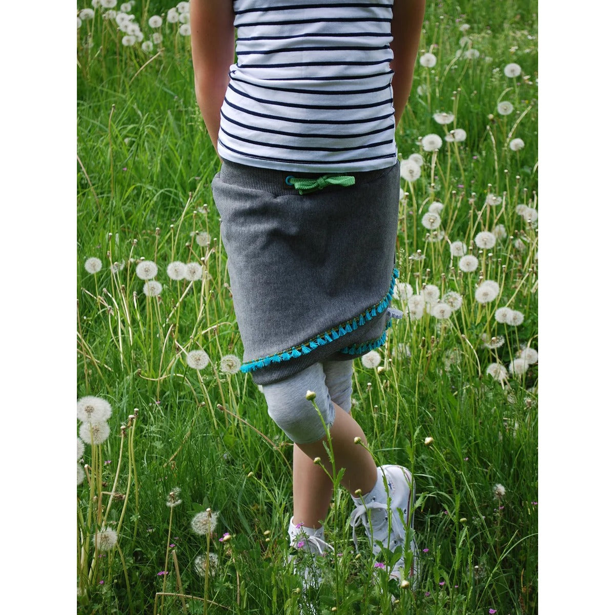Schnittmuster Shabby Skirt Kids – grauer Sweat mit grauem Bündchen und türkiser Fransenborte, Rückansicht auf einer Pusteblumenwiese