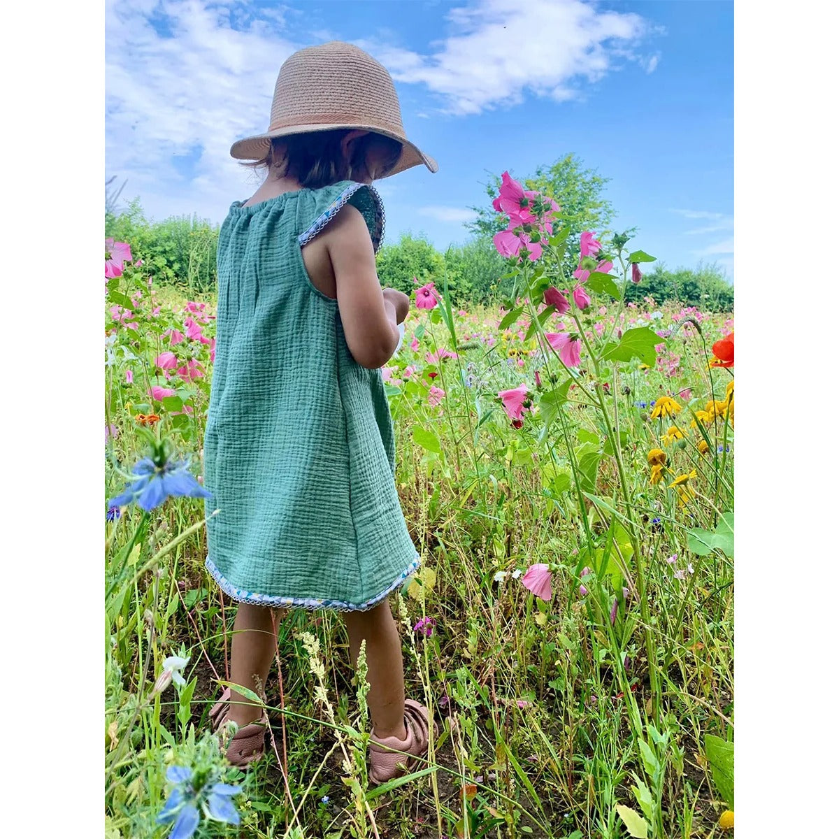 Schnittmuster Talia – zum Kleid verlängerte Variante aus grünem Musselin mit Flügelärmeln, Rückansicht auf einer Blumenwiese
