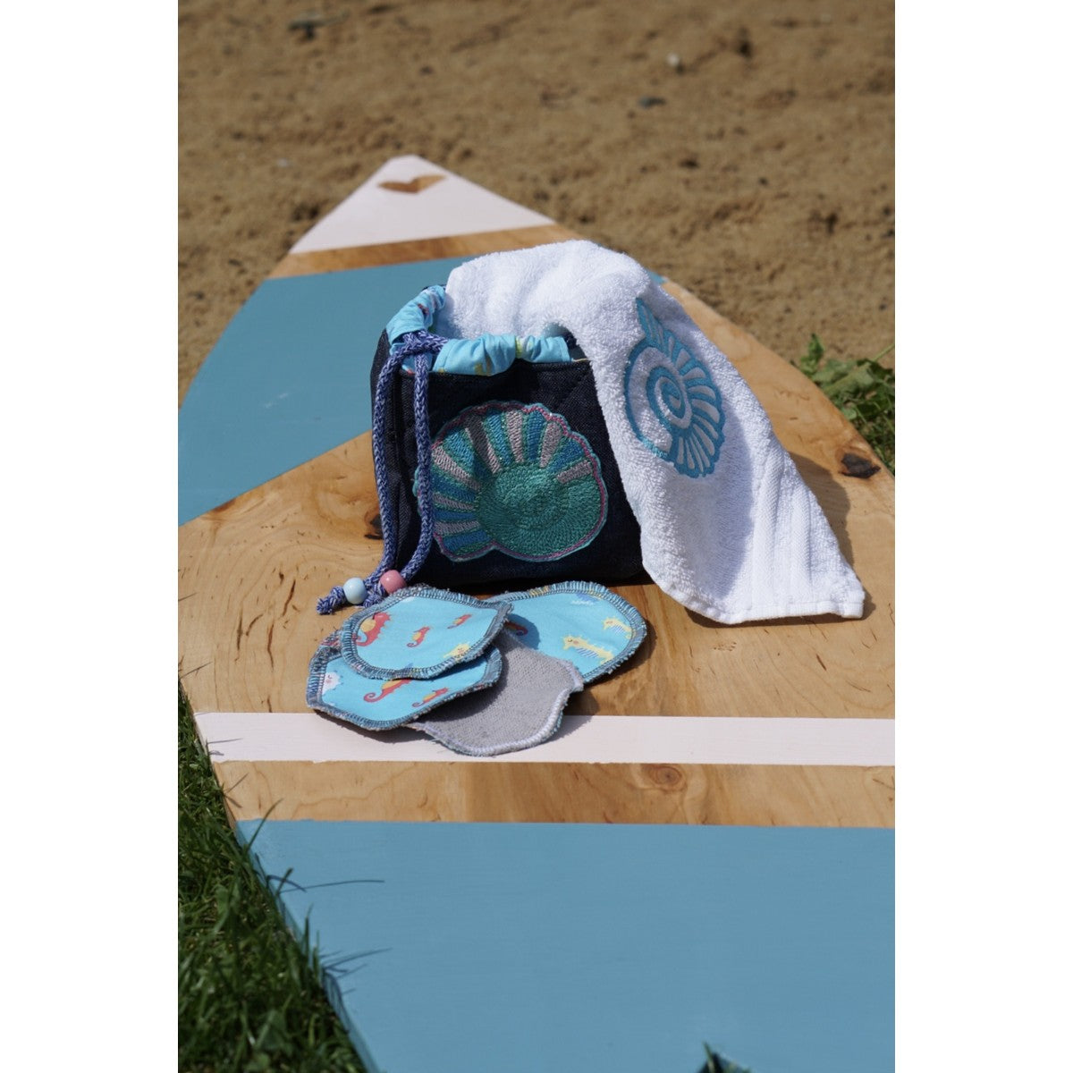 Stickdatei Frottee-Muschel – weißes Handtuch mit türkiser Muschel in einem Jeans-Utensilo mit gestickter Muschel, davor Kosmetikpads, fotografiert am Strand