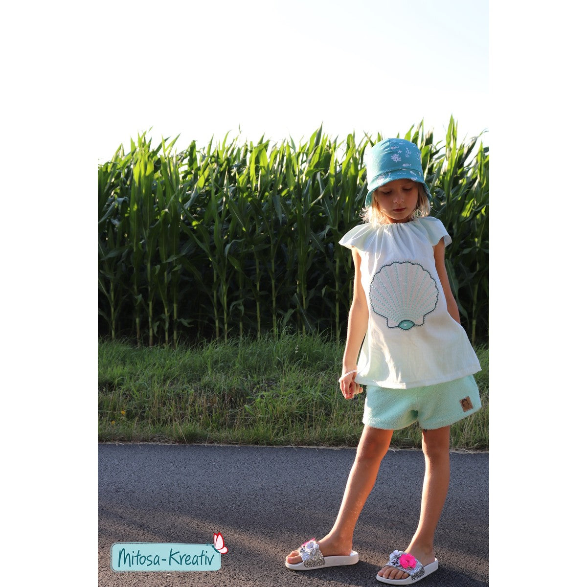 Stickdatei Muschel – Mädchen mit Sonnenhut im weißen Kleid mit großer Muschel und mintfarbenen Shorts vor einem Maisfeld