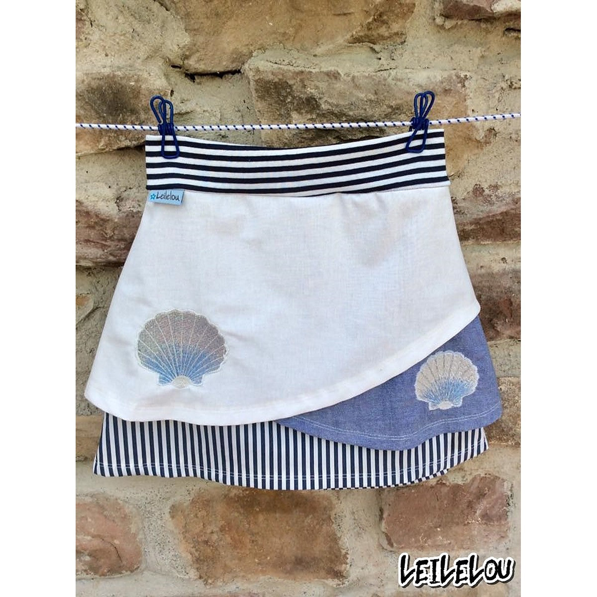 Stickdatei Muschel – weiß-blauer Wickelrock mit zwei gestickten Muscheln, an einer Leine vor einer Steinmauer